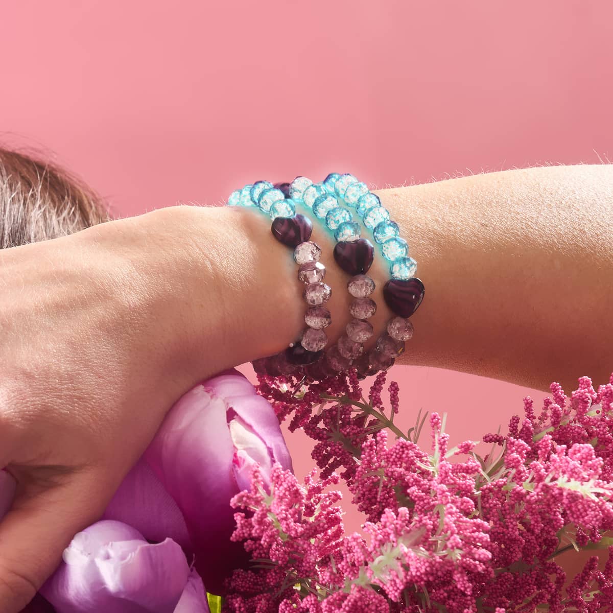 Plum glowing gradient hearts bracelets with blue and purple glass beads on clear elastic cords worn on wrist with pink flowers background by NOGU